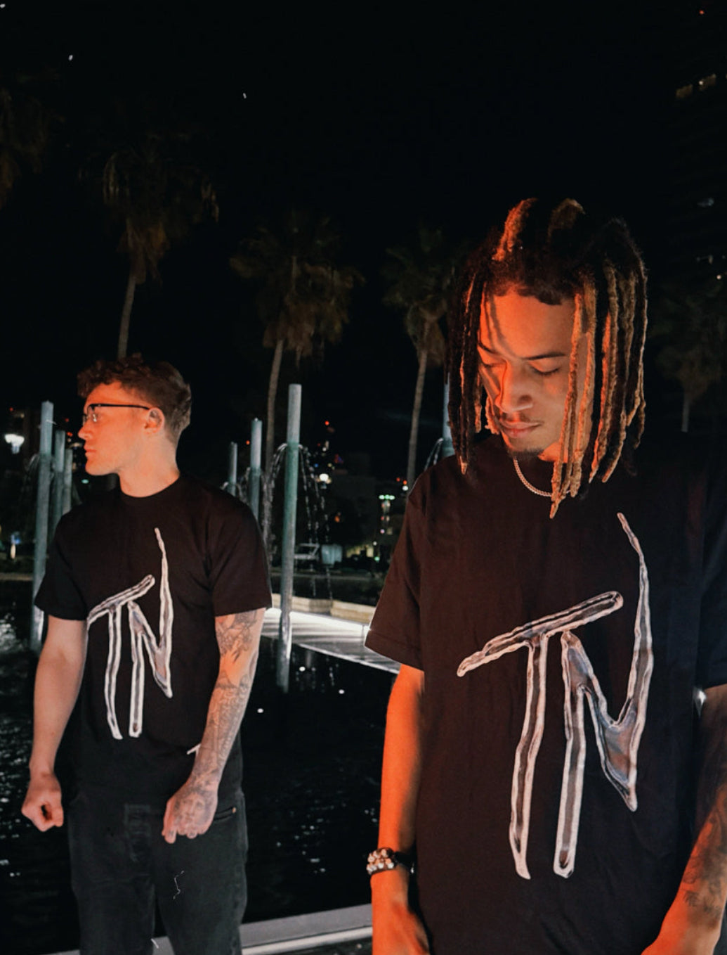 Two individuals wearing black t-shirts with a logo at night.